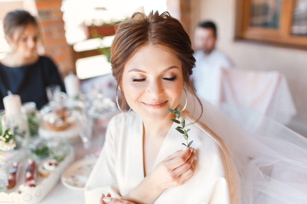 Morning portrait of the cute bride with branch. She sitting on the terrace and closed her eyes, touching her cheek with the branch of olive tree