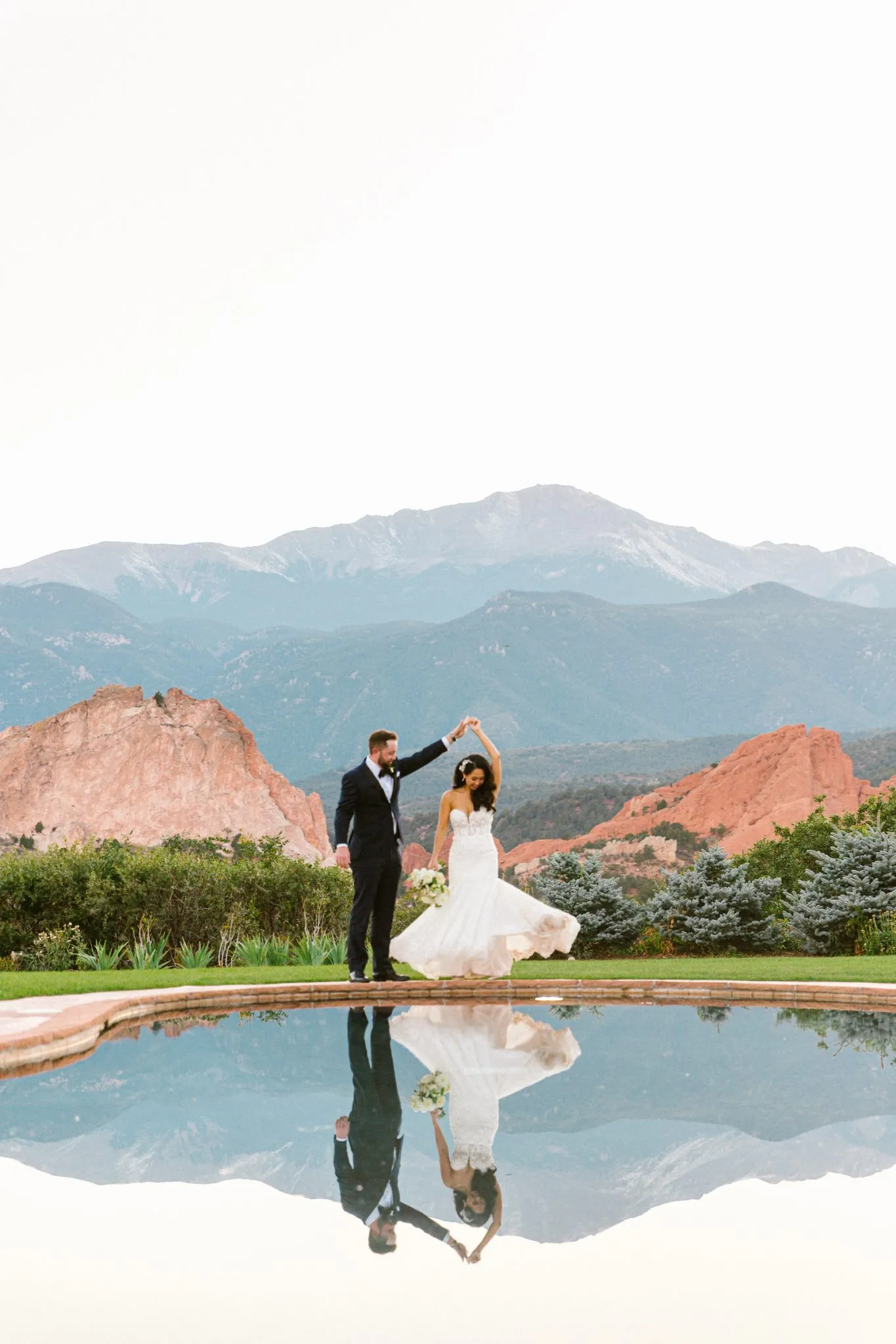 a couple in wedding attire standing by a pool with a mountain view