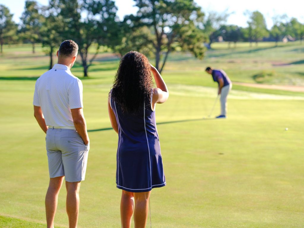 Two colleagues watching a teammate putt on a sunny golf course, perfect for outdoor company retreat ideas.