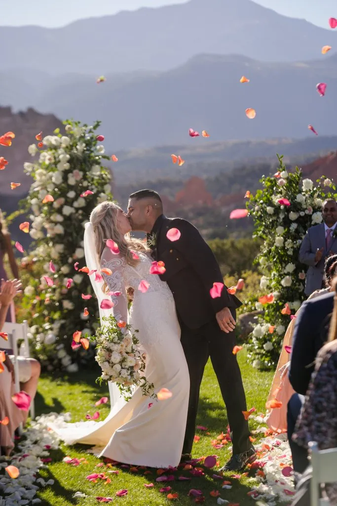 Newlyweds kissing beneath a floral arch during a luxury wedding ceremony with red rocks in the background.