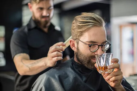 man in a barber chair having a drink while a barber gives him a hair cut