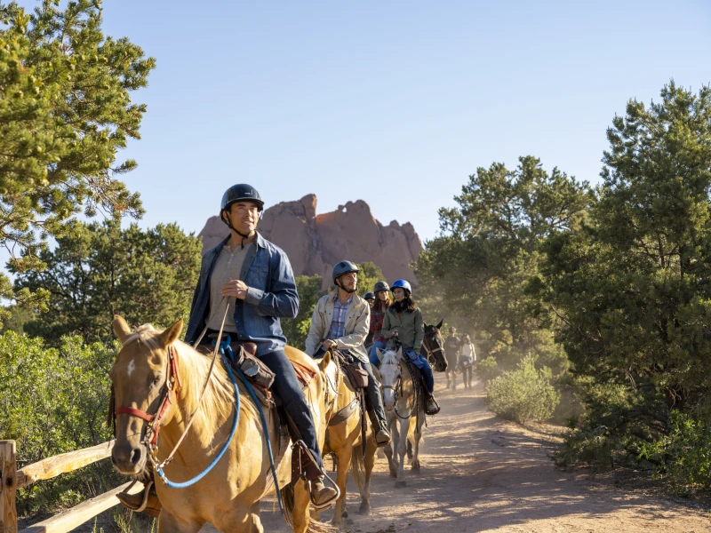 Group of people horseback riding on a dirt trail past red rock formations